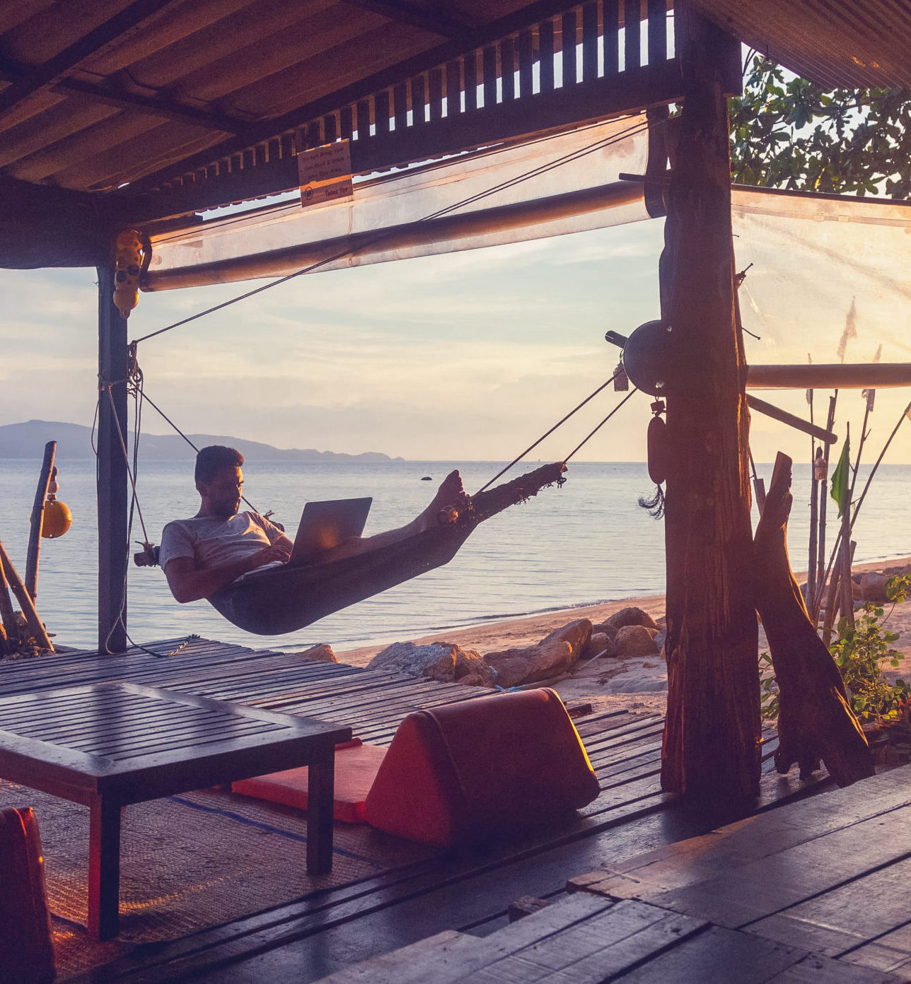 Young attractive guy a man with a beard in a hammock with a laptop on the background of the sea and sunset, distant work, freelancer, blogger, vacation and travel